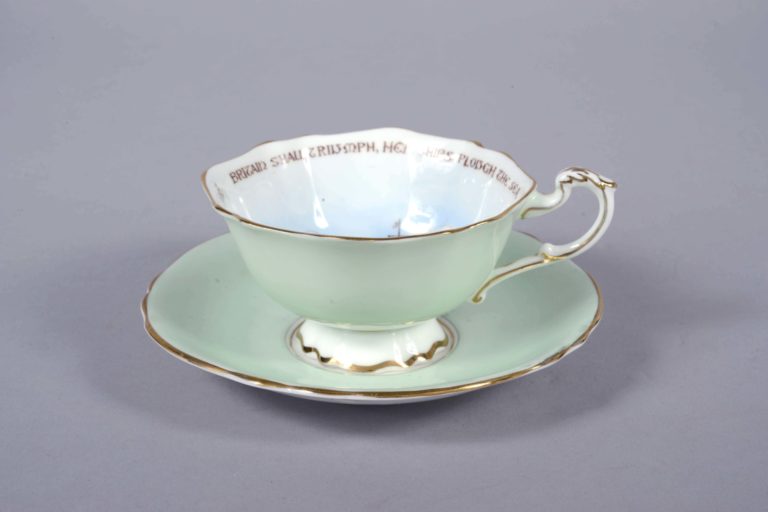 GEI-0007-Cup Green fine china cup with painting of three battleships. Words around the inside of the rim read: ‘Britain shall Triumph, her ships plough the sea, her standard be justice, her watchword be free’. Green fine china saucer with gold rim and Officers cap badge painted in the centre. This cup and saucer is part of the ‘Paragon Patriotic’ series produced during World War Two.