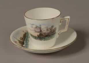 GEI-0023_GEI-0022-e1422843434422 Demi Tasse white porcelain coffee cup and saucer with a gold rim around the top. Front of cup is decorated with a picture of a World War One battleship firing a broadside with painted words: ‘H.M Battleship in action’. Top of saucer is decorated with a picture of a World War One torpedo boat with a white ensign at the jack. Words on the saucer read: ‘H.M Torpedo Boat’.