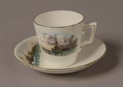 GEI-0023_GEI-0022-e1422843434422 Demi Tasse white porcelain coffee cup and saucer with a gold rim around the top. Front of cup is decorated with a picture of a World War One battleship firing a broadside with painted words: ‘H.M Battleship in action’. Top of saucer is decorated with a picture of a World War One torpedo boat with a white ensign at the jack. Words on the saucer read: ‘H.M Torpedo Boat’.